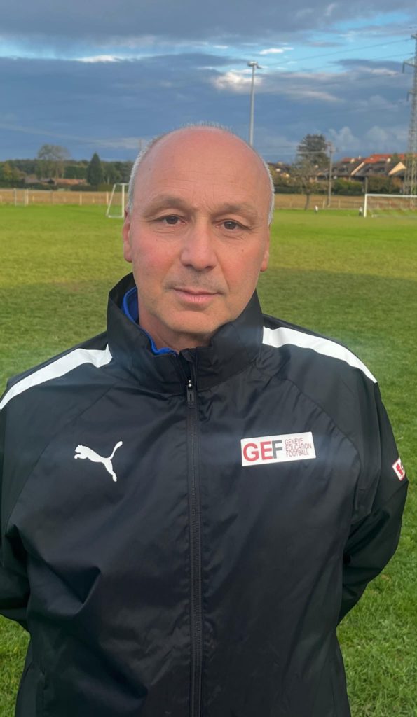 Portrait d'Antonio Antonazzo, scout expérimenté du programme Footeco, photographié sur un terrain de football à Genève. Il porte une veste aux couleurs du GEF, reflétant son rôle clé dans la détection des jeunes talents.
