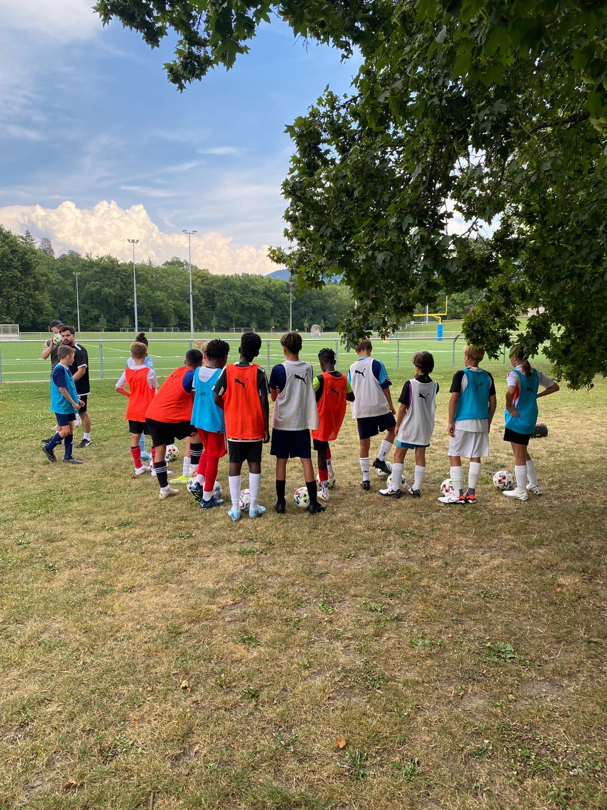 Un groupe de jeunes footballeurs, portant des chasubles colorés, écoute attentivement les consignes de leurs entraîneurs à l'ombre d'un arbre pendant une séance d'entraînement du programme FootEco à Genève.