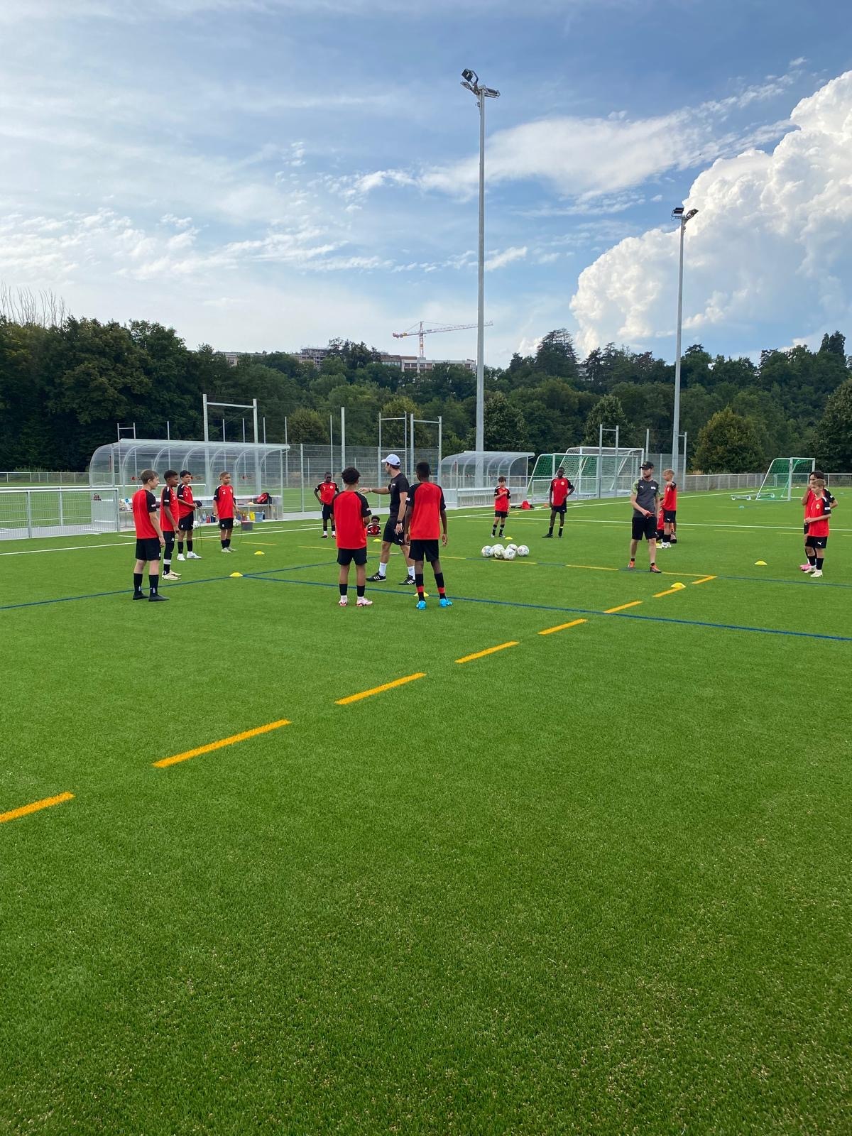 Séance d'entraînement FootEco à Genève : de jeunes footballeurs en maillots rouges perfectionnent leurs techniques sur terrain synthétique, encadrés par des coachs expérimentés.