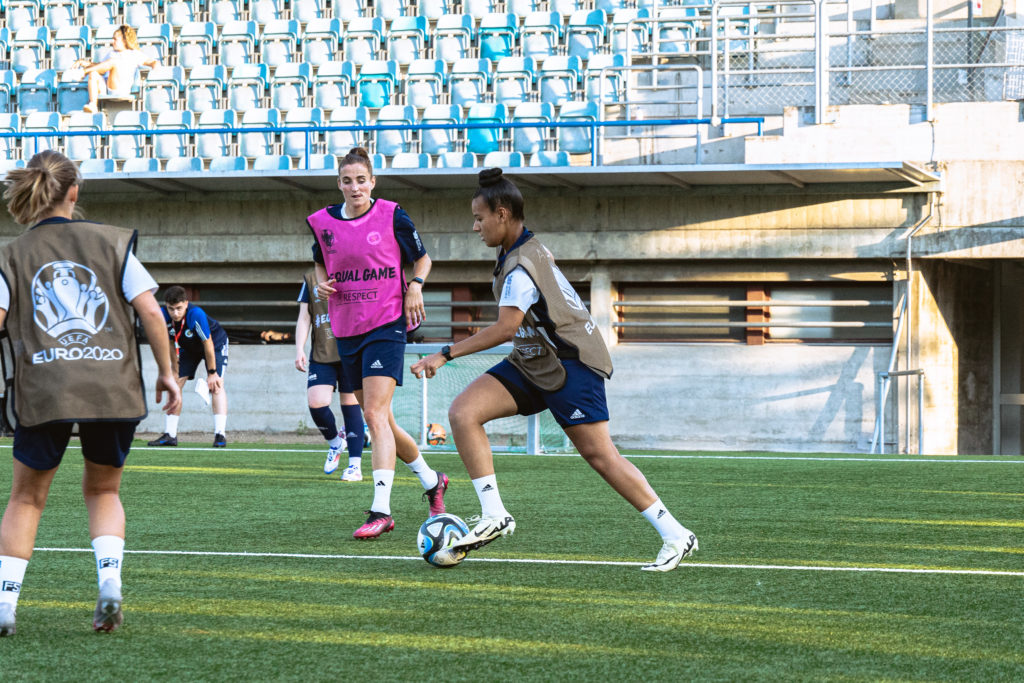 Entraînement de football féminin au stade avec Leila Marrel, jeune entraîneure et joueuse du GEF, en action avec d'autres joueuses sur le terrain.
