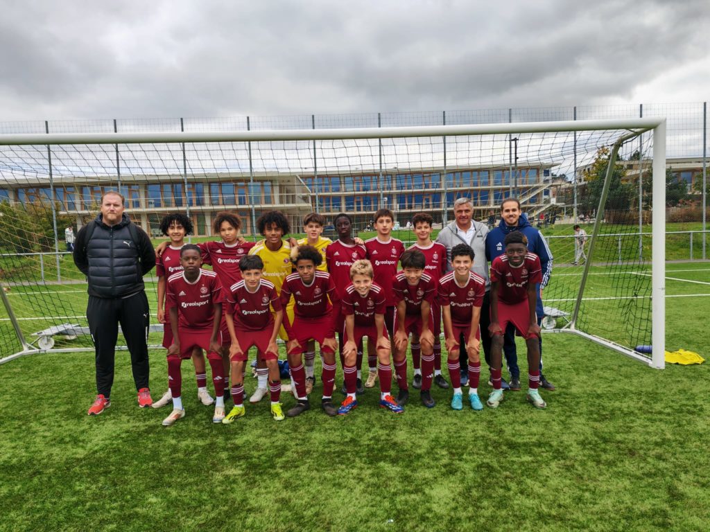 Photo de l'équipe FE13 et FE14 du Genève Education Football (GEF) posant devant un but sur un terrain de football. Les jeunes joueurs portent un maillot rouge et sont accompagnés de leurs entraîneurs, sous un ciel nuageux
