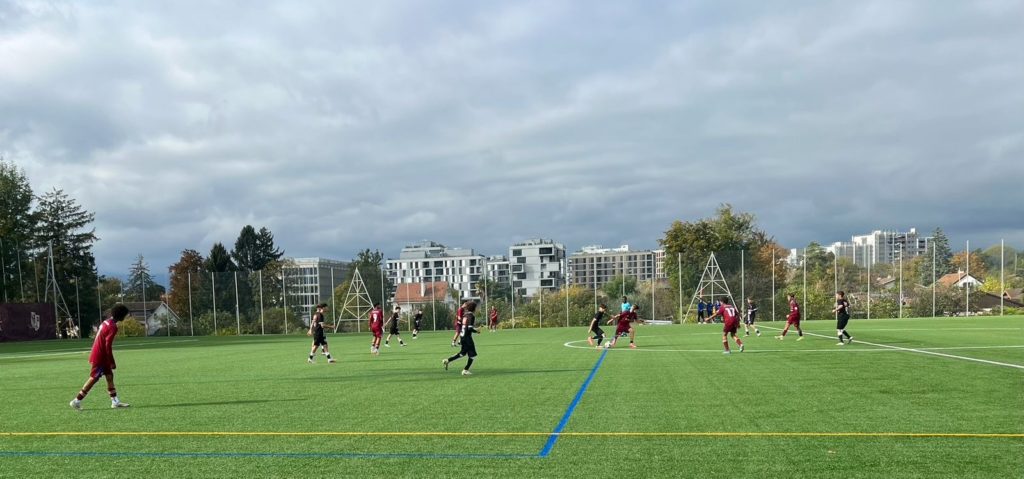 Opposition Footeco sur synthétique à Genève : maillots grenat contre noirs, large vue du terrain.